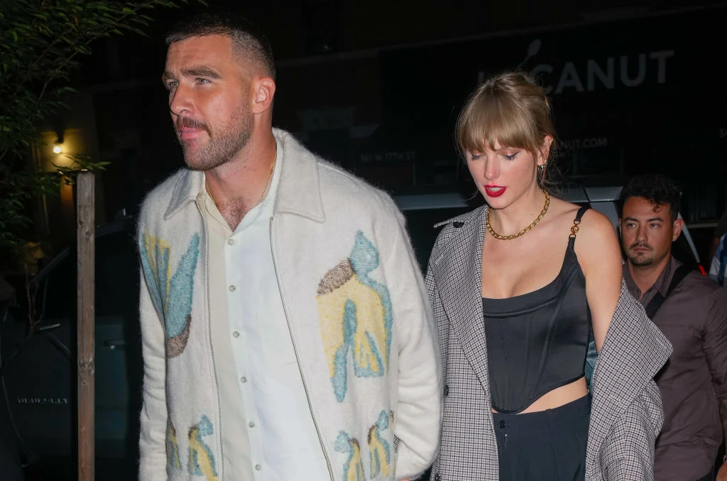 Taylor Swift walking beside a man outside at night, with both dressed in casual outfits.
