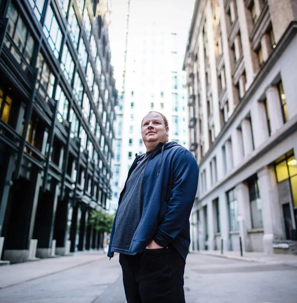 Parker Conrad standing on a city street between tall office buildings, wearing a dark blue jacket and black pants.